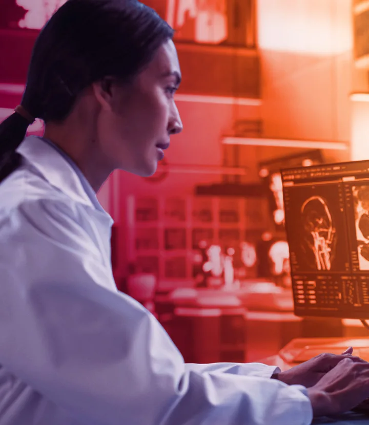 A female scientist in a white lab coat sits at a desk in a red‑lit laboratory, intently viewing a computer screen showing detailed brain scan images; a simple white neuron icon is overlaid in the lower left corner.
