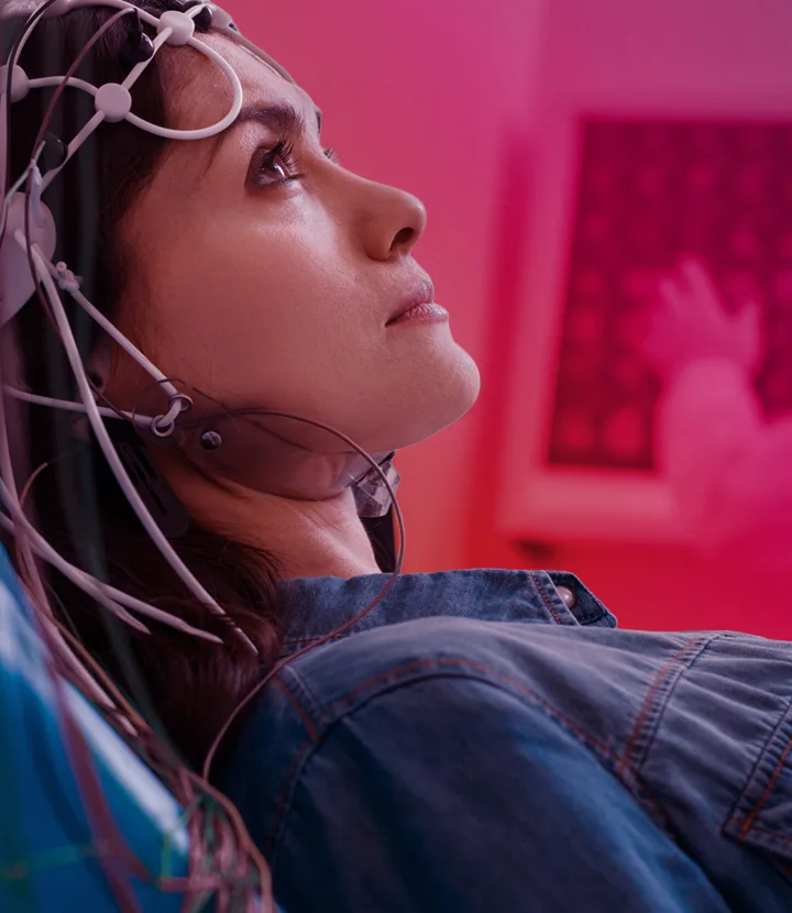 A side view of a young woman reclining with an array of EEG electrode sensors and wires attached to her head, gazing upward against a pink‑purple tinted background with a simple white microchip/circuit icon overlaid in the upper right.