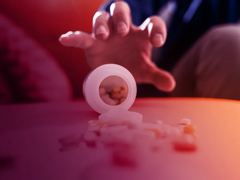 Hand reaching for spilled prescription pills from an open pill bottle, representing substance use or medication misuse.