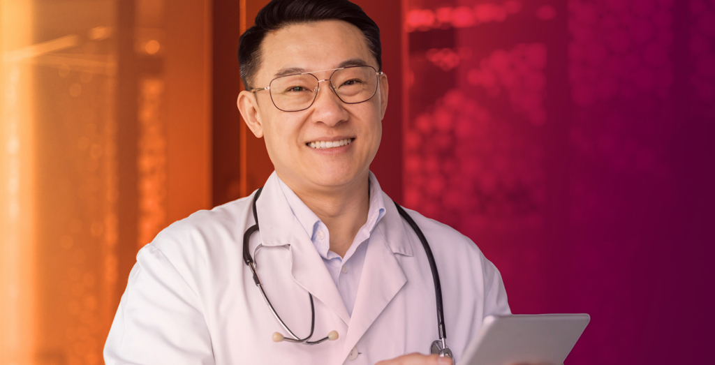 Smiling male doctor wearing glasses and a white coat with a stethoscope, holding a tablet against a warm orange-to-purple gradient background.