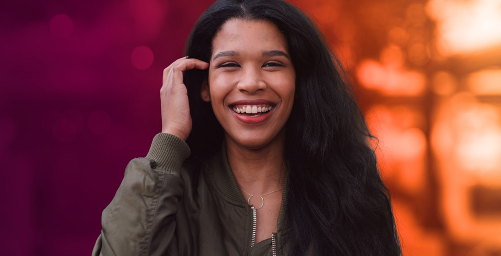 Young woman with long dark hair and a green jacket smiling broadly, one hand near her hair, set against a blurred orange-and-purple gradient background with bokeh light effects.