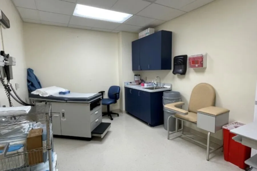 Clinical exam room with exam table, blue cabinets, sink, and phlebotomy chair.