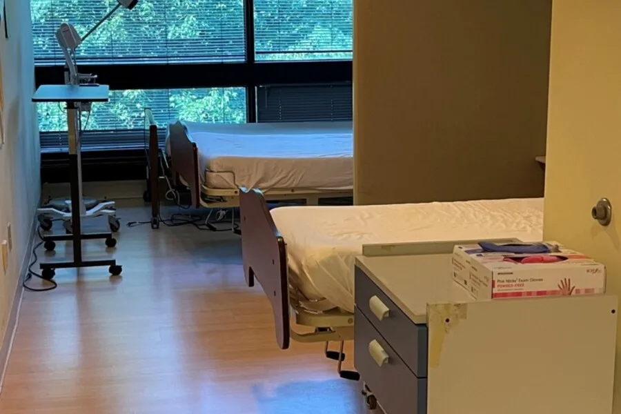 Two-bed patient room with nightstand and supplies, windows along back wall.