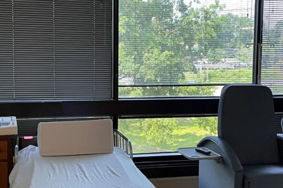 Patient bed and recliner beside large windows with blinds and green view.