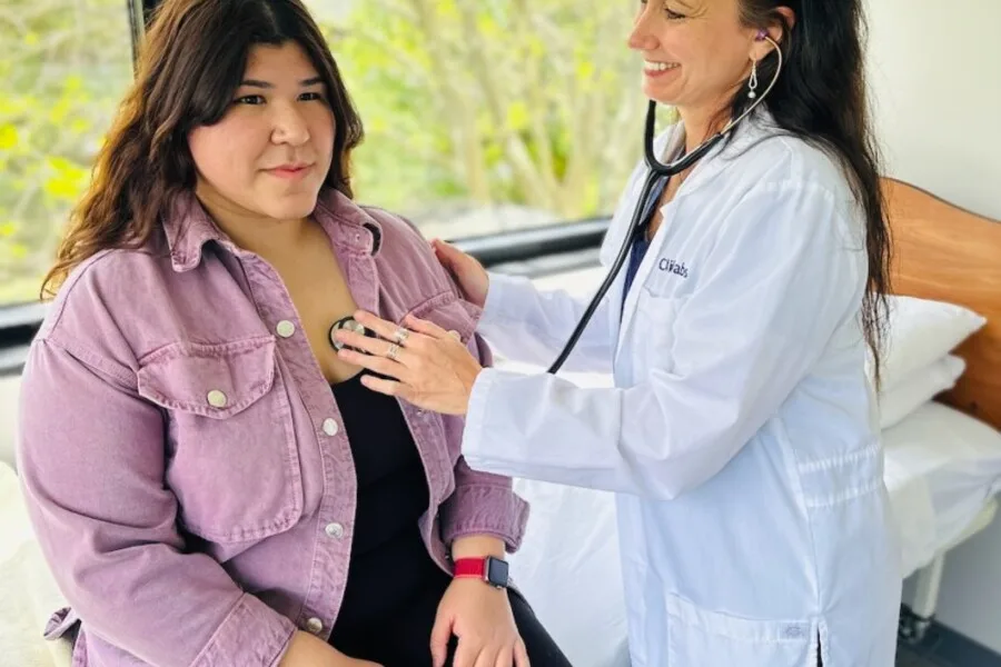 Clinician using a stethoscope to listen to a patient’s chest beside an exam bed.”