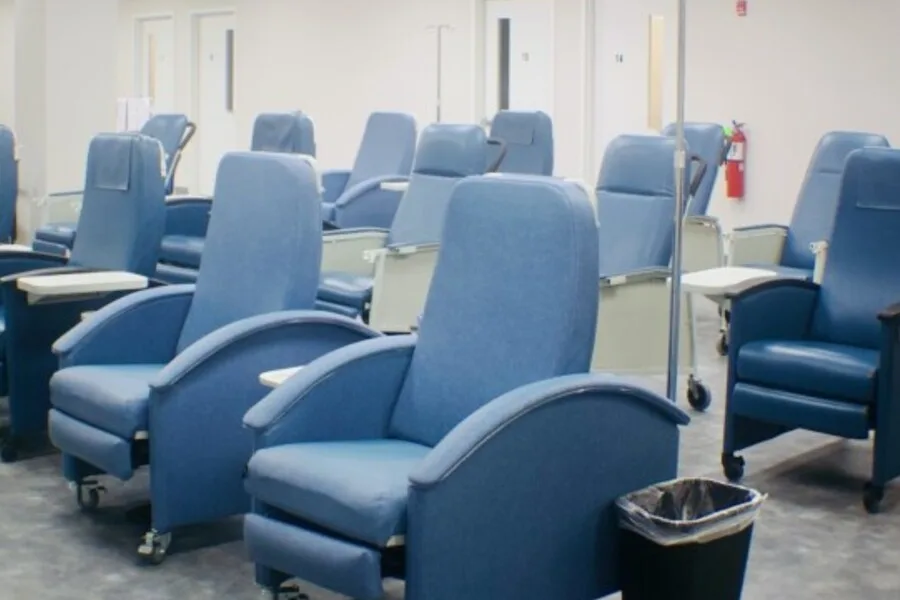 Large treatment room with rows of blue recliner chairs, tray tables, and IV poles.