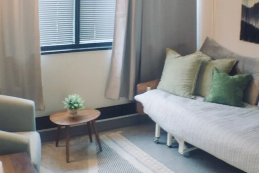 Cozy patient room with daybed, green pillows, small round table, and window with curtains.