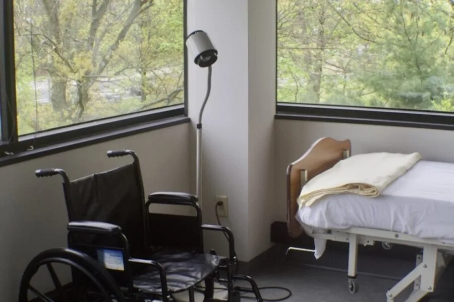 Corner of a hospital room with wheelchair beside a bed by large windows overlooking trees.”