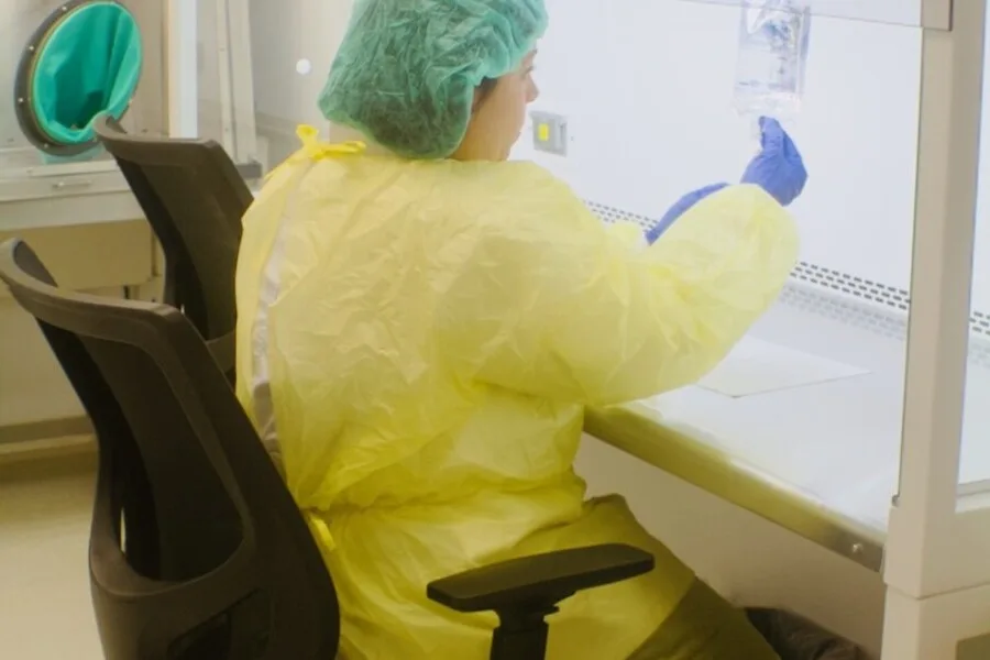 Technician in gown, gloves, and cap preparing medication inside a sterile biosafety hood.