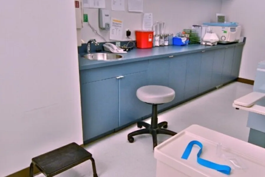 Clinical exam room with blue cabinets, sink, stool, and medical supplies.