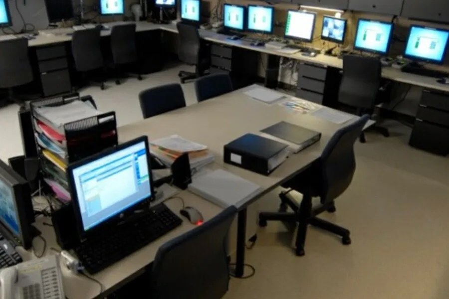 Monitoring room with rows of computers at workstations around a central table.
